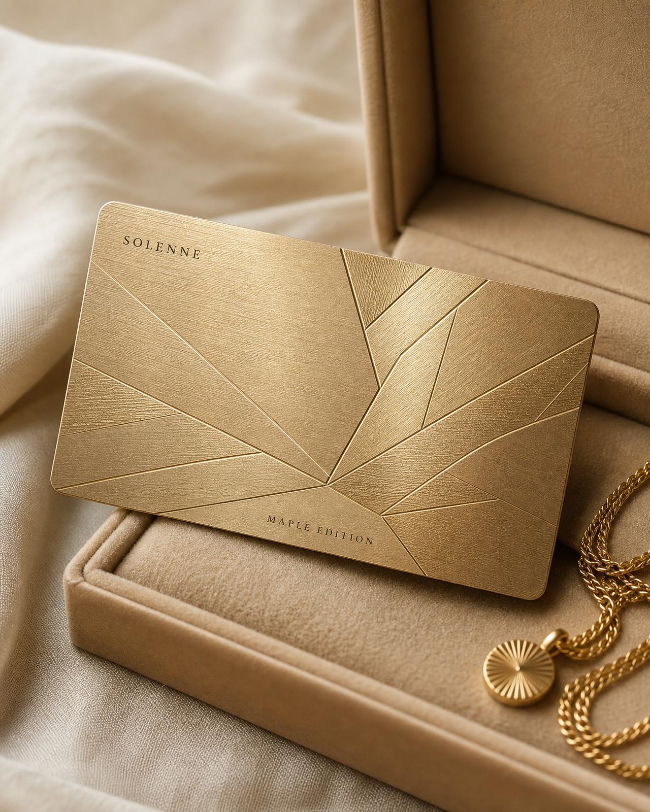 Maple Edition brushed champagne gold card with engraved geometric veining, half lifted from a sand suede box