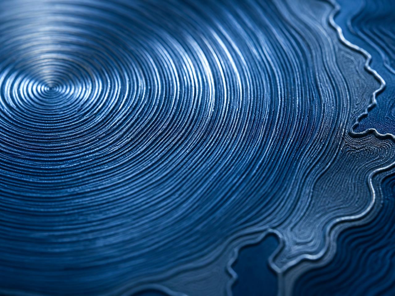 Macro detail of the Pacific Edition surface — engraved concentric wave rings and coastal contours in deep blue titanium